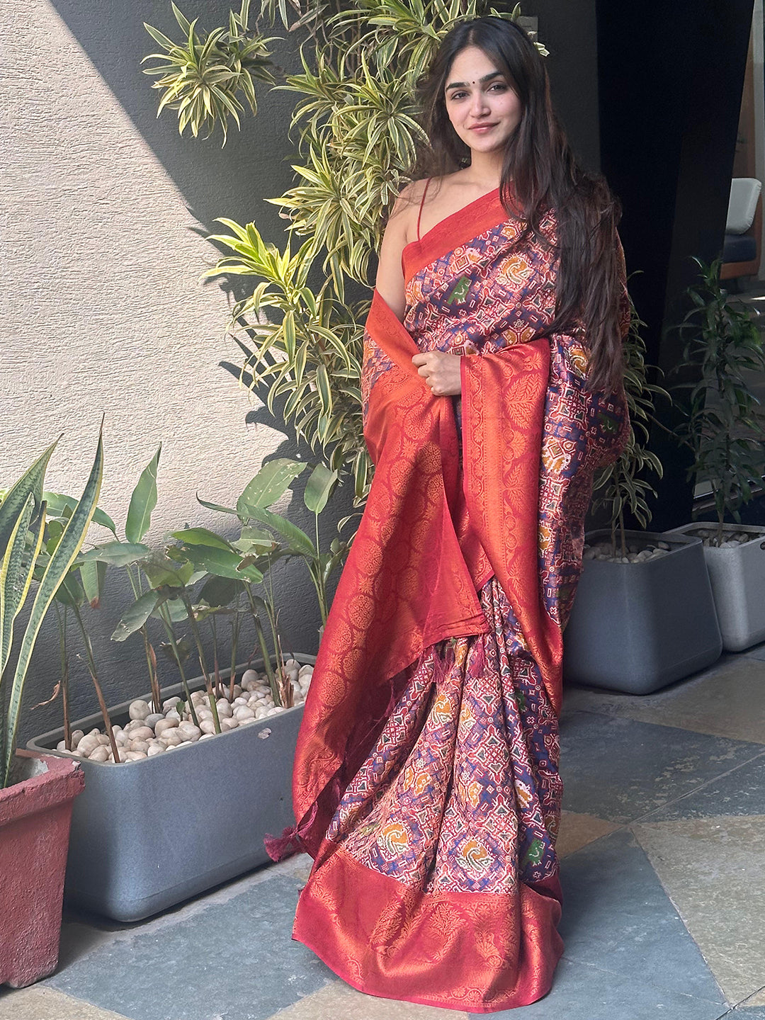 Woman in red Patola saree with vibrant patterns, standing indoors.