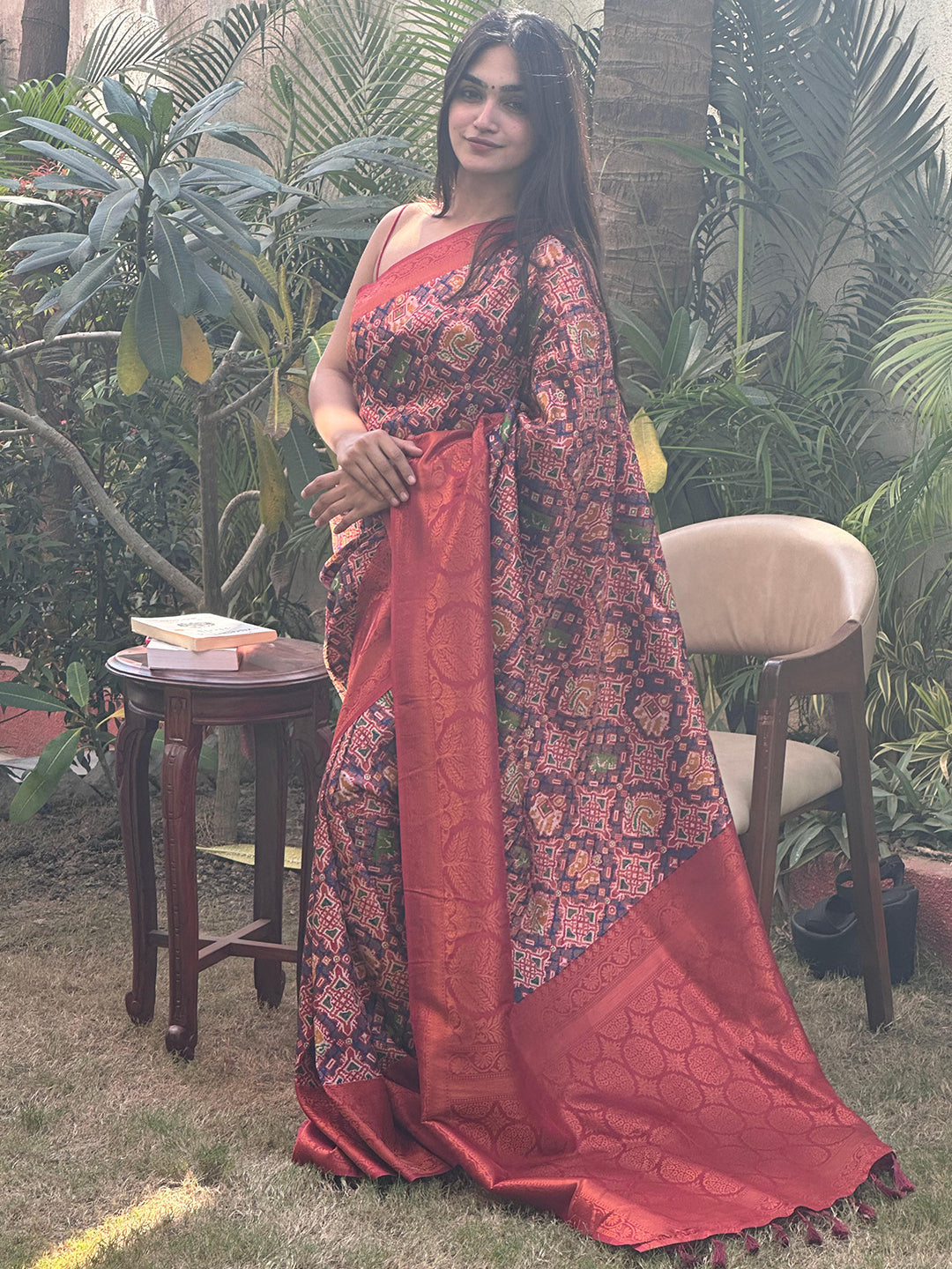 Woman standing in red Patola saree with complex patterns, garden setting.