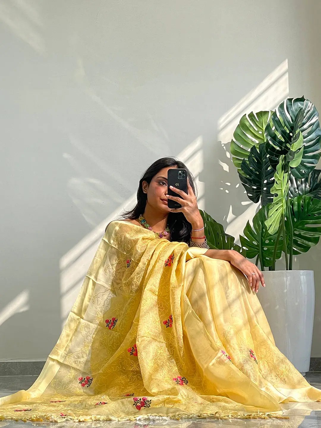 Model seated in yellow saree with floral embroidery, holding phone.