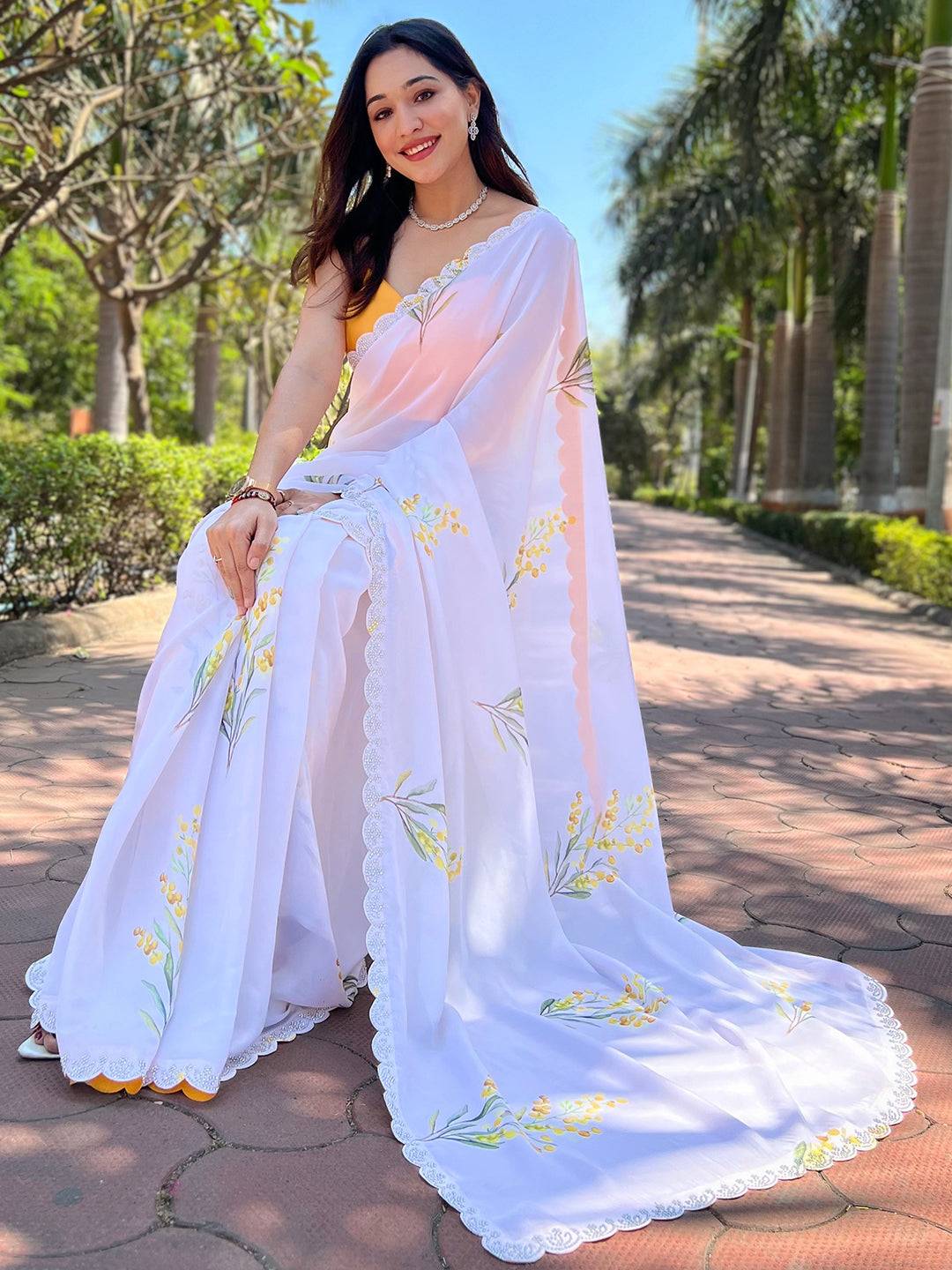 Seated woman in white floral satin georgette saree with zircon border